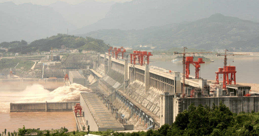 A panoramic view of the Three Gorges Dam in China, showcasing its massive concrete structure spanning the Yangtze River, with lush green hills in the background.